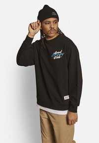 Schwarzer Sweatshirt mit einem farbenfrohen "Good Company Club"-Logo, gerippten Bündchen und einem Etikett am Saum. Trägt eine schwarze Strickmütze.