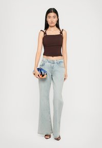 Bruine, aansluitende crop top met verstelbare bandjes, gecombineerd met lichtblauwe flared jeans. Het model houdt een kleurrijke abstracte handtas vast.