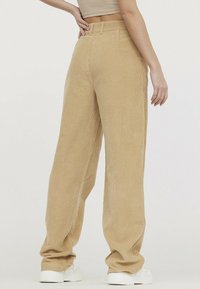 Pantalons évasés en velours côtelé beige clair, avec une texture côtelée verticale, taille haute et coupe ample ; associés à des baskets blanches.