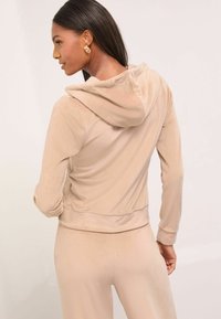 Sudadera de terciopelo color marrón claro con capucha ajustable, puños y cinturilla ajustados. Textura suave con un acabado blando, combinada con pantalones a juego.