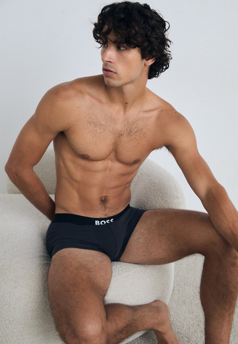 Schwarze Boxer-Briefs für Herren mit einem breiten elastischen Bund, auf dem "BOSS" steht, liegen auf einer hellen, strukturierten Oberfläche. Natürlicher Hautton sichtbar.