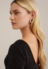 Juoda suknelė su blizgiu tekstūru, giliu V formos nugarėlė ir prigludusios rankovės. Modelis dėvi blizgius sidabrinius ašarų formos auskarus. Ilgi, banguoti šviesūs plaukai.