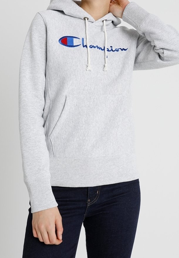 Personne portant un sweat à capuche Champion gris clair avec un logo bleu et rouge, jean foncé, une main touchant le cou et l'autre détendue sur le côté.