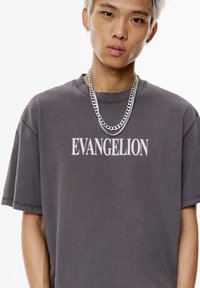 Harmaa ylisuuri t-paita, jossa on valkoiset "EVANGELION"-tekstit. Käyttäjällä on hopeaketju kaulassa ja näkyvät korvapaikat.
