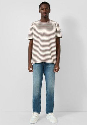 Jonge man staand, gekleed in een bruin-wit gestreept T-shirt met korte mouwen, blauwe spijkerbroek en witte sneakers, tegen een effen lichte muur.