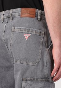 Sivek denim cargo kratke hlače s rjavim usnjenim oznako in rdečo-belo trikotno oznako na zadnjem žepu. Vidne so podrobne šive.