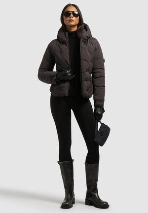 Un mannequin porte une doudoune sombre, une tenue noire, des gants, des lunettes de soleil et des bottes montantes jusqu’aux genoux, et tient un petit sac à bandoulière sur un fond uni.
