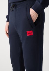 Marineblauwe sweatpants van zachte stof hebben zijzakken en een rood rechthoekig logo-label met de tekst "HUGO" op de dij.