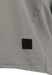 Grau Baumwoll-T-Shirt-Stoff mit glatter Textur; verfügt über ein kleines schwarzes Stoff-Logo-Patch auf der unteren linken Seite. Der Saum ist leicht abgerundet.