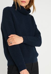 Pull en mailles bleu marine avec un motif texturé, col roulé haut et poignets et ourlet côtelés. Matière douce, coupe décontractée, design légèrement oversize.