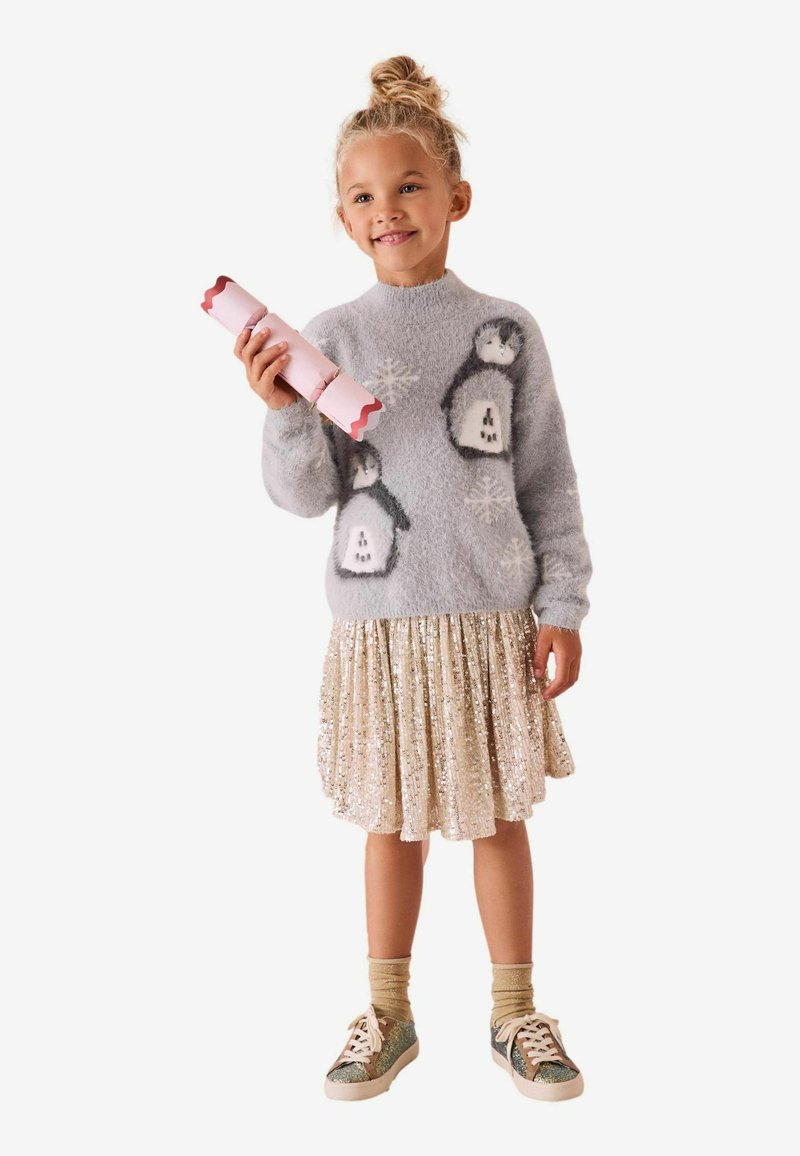 Maglione grigio morbido con design di pinguini, gonna plissettata di paillettes oro chiaro, calze dorate e sneakers glitterate, mentre tiene un cracker da party rosa.