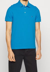 Muž v jasně modrém polo tričku s krátkým rukávem, se třemi knoflíky a malým logem na hrudi, v kombinaci s černými kalhotami.