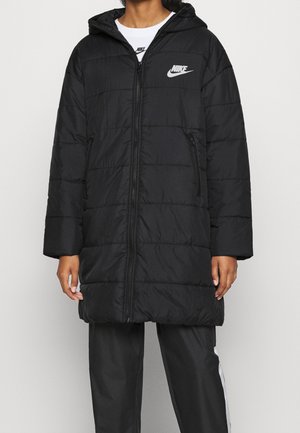 Person trägt eine schwarze, gesteppte Nike-Kapuzensjacke mit vorderem Reißverschluss und Reißverschlusstaschen, kombiniert mit schwarzen Hosen mit weißen Seitenstreifen.