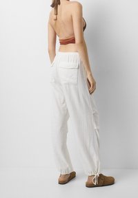 Pantalones de lino blancos con puños ajustables, que cuentan con dos bolsillos traseros, combinados con una blusa marrón y zapatos marrones sin cordones.