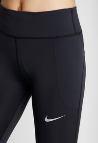 Close-up van de taille en bovenbenen in zwarte Nike leggings met een zijzak en het witte Nike swoosh-logo op de dij.