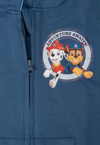 Felpa con zip blu che presenta una grafica circolare di due cani dei cartoni animati, uno con un cappello da pompiere rosso e l'altro con un cappello da poliziotto blu.