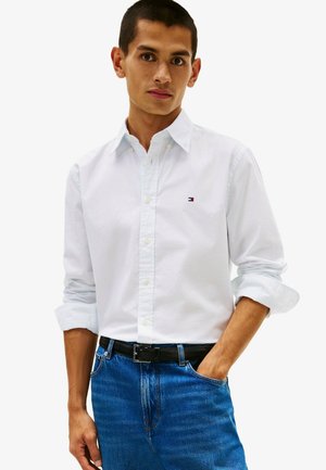 Jeune homme portant une chemise blanche boutonnée avec manches retroussées et un jean bleu, posant avec une main dans la poche sur un fond uni.