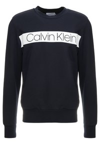 Námořnické modré mikina vyrobená z měkké tkaniny, s bílým horizontálním pruhem s textem "Calvin Klein". Hladké manžety a lem.