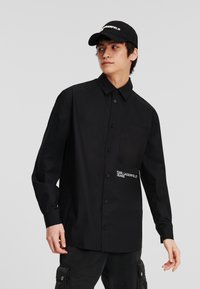 Juodas marškiniai su užsegimu, turintys krūtinės kišenę, puošti balta siuvinėta "KARL LAGERFELD JEANS" užrašu. Suderinti su juodomis cargo kelnėmis ir kepure.