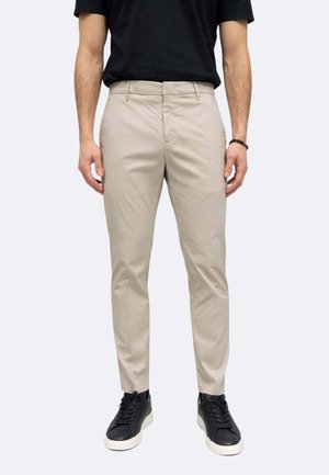 Uomo che indossa pantaloni beige slim fit, maglietta nera, sneakers nere con suola bianca, in piedi contro uno sfondo chiaro uniforme.