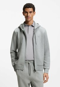 Felpa con cappuccio grigia con cerniera, cappuccio con coulisse, tessuto morbido e tasche laterali. Indossata sopra una polo grigio chiaro e pantaloni abbinati.