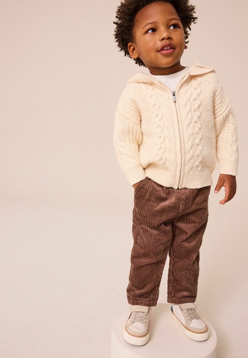 Suéter de punto trenzado crema con cremallera, pantalones de pana marrones con rayas verticales y zapatillas blancas con detalles en marrón. De pie sobre un pedestal.