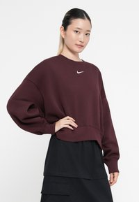 Μαρούν φούτερ με στρογγυλή λαιμόκοψη και χαμηλούς ώμους, με λευκό logo της Nike. Φορεμένο πάνω από μια μαύρη πολυεπίπεδη φούστα. Μαλακή υφή.