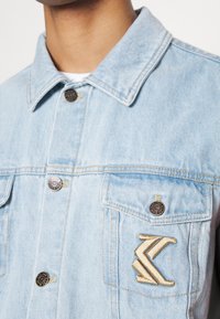 Light blue denim jacket featuring a pointed collar, two front pockets, gold embroidery detail, and black metal button accents.