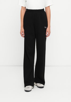 Person iført sorte bukser med brede ben og elastisk talje, hvid cropped top og grå Nike sneakers, står mod hvid baggrund.