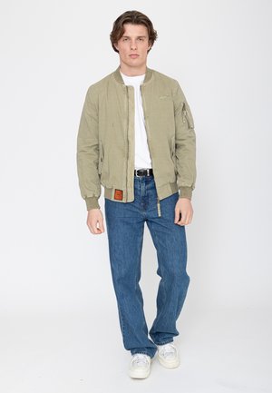 Young man standing wearing beige bomber jacket, white t-shirt, blue jeans, white sneakers, on plain white background.