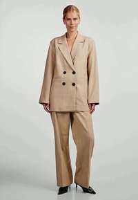 Beige double-breasted blazer with black buttons, wide sleeves, and a relaxed fit, paired with matching wide-leg trousers and black heels.