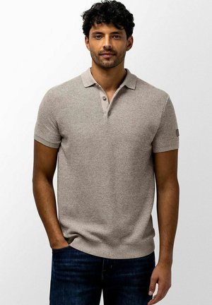 Man with short curly black hair wearing a beige short-sleeve knit polo shirt and dark blue jeans standing against a plain background.