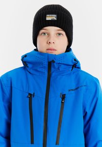 Blaue wasserdichte Jacke mit hohem Kragen, schwarzen Reißverschlüssen und Belüftungsöffnungen. Kombiniert mit einer schwarzen Rippmütze mit einem Logopatch.