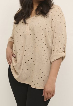 Vrouw die een beige blouse met lange mouwen en zwarte polka dots draagt, gecombineerd met een zwarte broek, poseert met één hand op haar heup en één arm ontspannen.