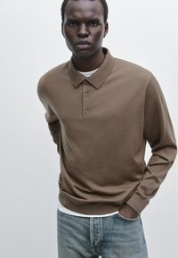 Brown long-sleeve polo shirt with a collar, three buttons, and a textured knit. Paired with light blue distressed jeans.