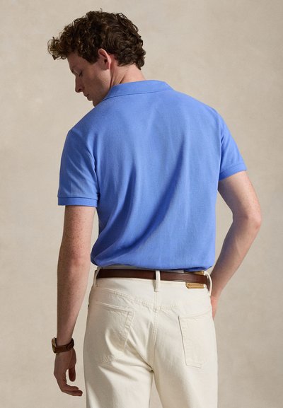 Man met krullend haar die een blauw poloshirt met korte mouwen, een witte broek, een bruine riem en een bruine polshorloge draagt, staand tegen een effen achtergrond.
