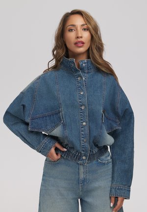 Kobieta w jeansowej kurtce z wysokim kołnierzem, zapinanej na guziki, krótkiej z elastycznym paskiem w talii, oraz w wysokich jeansach, pozująca z jedną ręką w kieszeni.