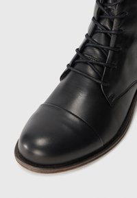 Botín de cuero negro hasta el tobillo con puntera redonda, sutil detalle de costura y cordones resistentes. Presenta una textura suave y suela de color marrón en contraste.