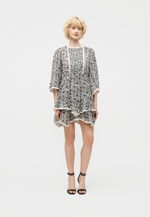 Mujer con cabello corto y rizado rubio, vestida con un vestido de estampado floral en blanco y negro y zapatos de tacón con tiras en el tobillo, posando contra un fondo liso.