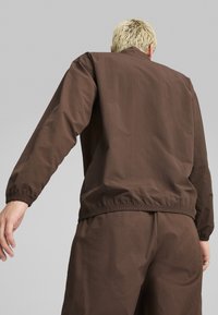 Brun lätt jacka med elastiska ärmslut och nederkant, med en slät yta och minimal design, bärs med matchande byxor.