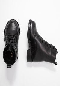 Botas de tobillo de cuero negro con punta redonda, cordones en la parte delantera, detalles cosidos y un tacón bajo. Presenta una lengüeta en la parte trasera.