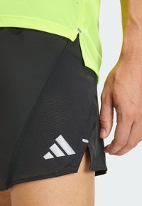 Close-up of a person wearing black athletic shorts with a white Adidas logo and a neon yellow shirt, showing their right hand relaxed by their side.