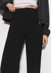 Gros plan sur une personne portant un pantalon noir taille haute, un haut blanc raccourci, une veste noire zippée, avec des bagues décoratives à la main gauche.