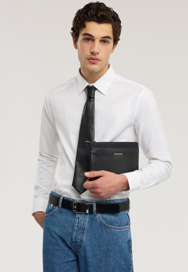 Giovane uomo con camicia bianca e cravatta nera tiene una borsa in pelle nera, indossa jeans blu e cintura nera su sfondo semplice.