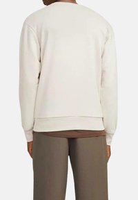 Sudadera beige con cuello redondo y puños acanalados, con un corte relajado y una tela suave, combinada con pantalones marrones.