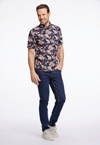 Camicia a maniche corte floreale con sfondo blu scuro e fiori rossi e bianchi, abbinata a jeans blu scuri e sneakers chiare.