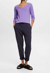 Fioletowy sweter w serek zestawiony z ciemnoszarymi spodniami dresowymi. Czarne sandały z akcentami w postaci kokardek. Casualowy, miękki materiał z luźnym krojem.