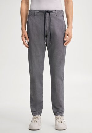Homme portant un pantalon gris à cordon et des baskets blanches, debout devant un fond uni.