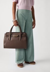 Borsa a tracolla in pelle marrone con design strutturato, dettaglio del logo dorato e doppi manici. Abbinata a pantaloni in corduroy verde chiaro a gamba larga e scarpe marroni.