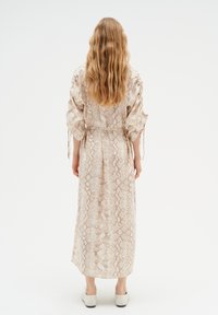 Pitkä, käärmeennahkakuosisessa pehmeässä beigen värisessä aarella, joka on varustettu vyöllä, leveillä hihoilla ja virtaavalla muotoilulla, jota täydentää hienovarainen tekstuuri.